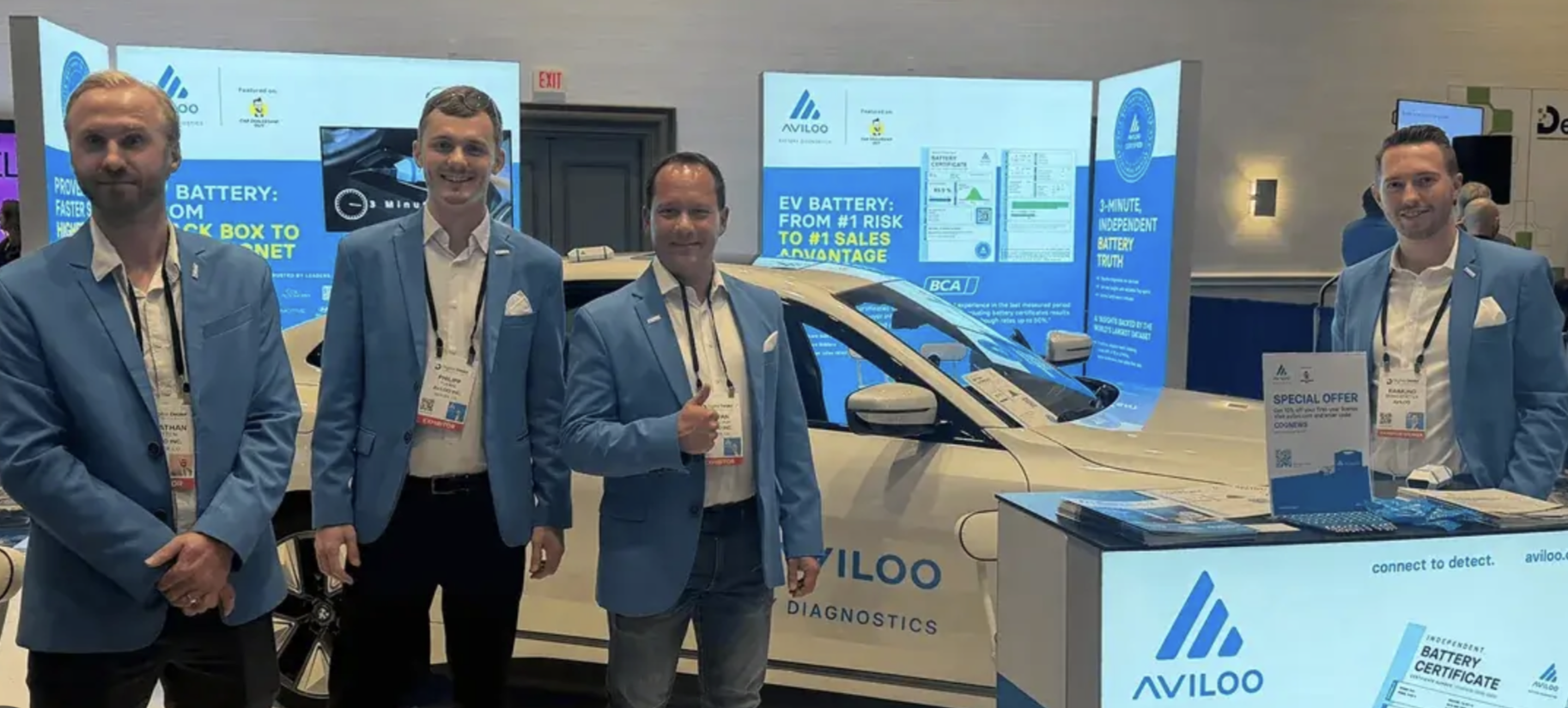Four AVILOO team members in blue blazers standing at the AVILOO booth beside a white electric car
