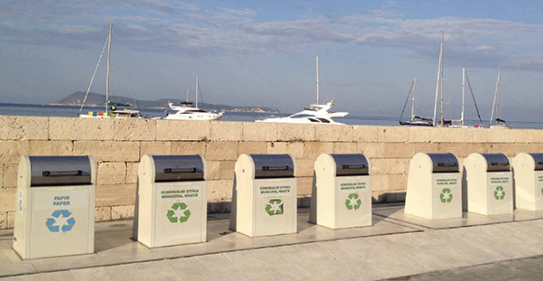 Row of white EUROTIM underground waste containers along a harbor promenade with boats in the background