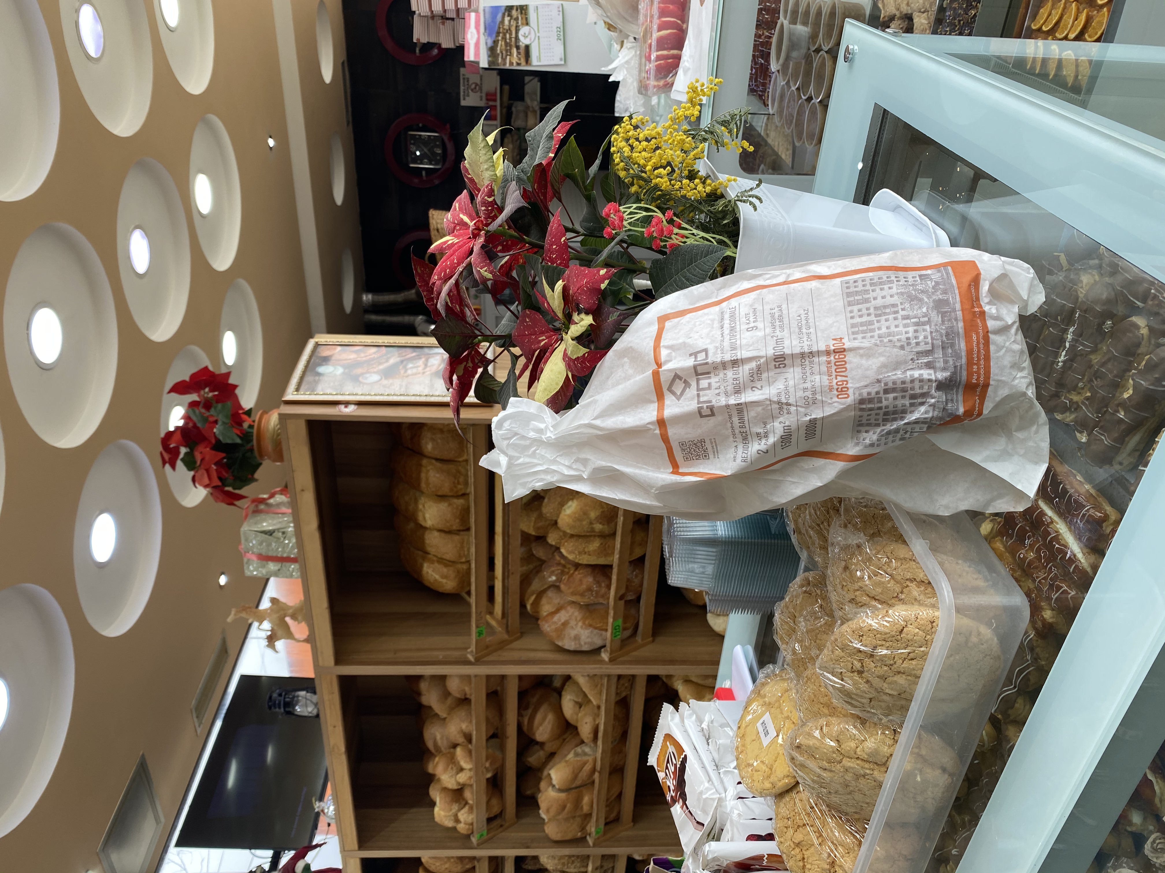 A white paper bread bag printed with a Grand Gallery advertisement, standing on a bakery counter next to a poinsettia plant